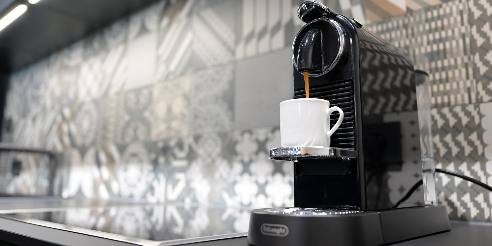 Schwarze Espressomaschine mit weißer Tasse, aus der Kaffee in die Tasse fließt, vor einer Wand mit grauem, gemustertem Fliesenmuster.